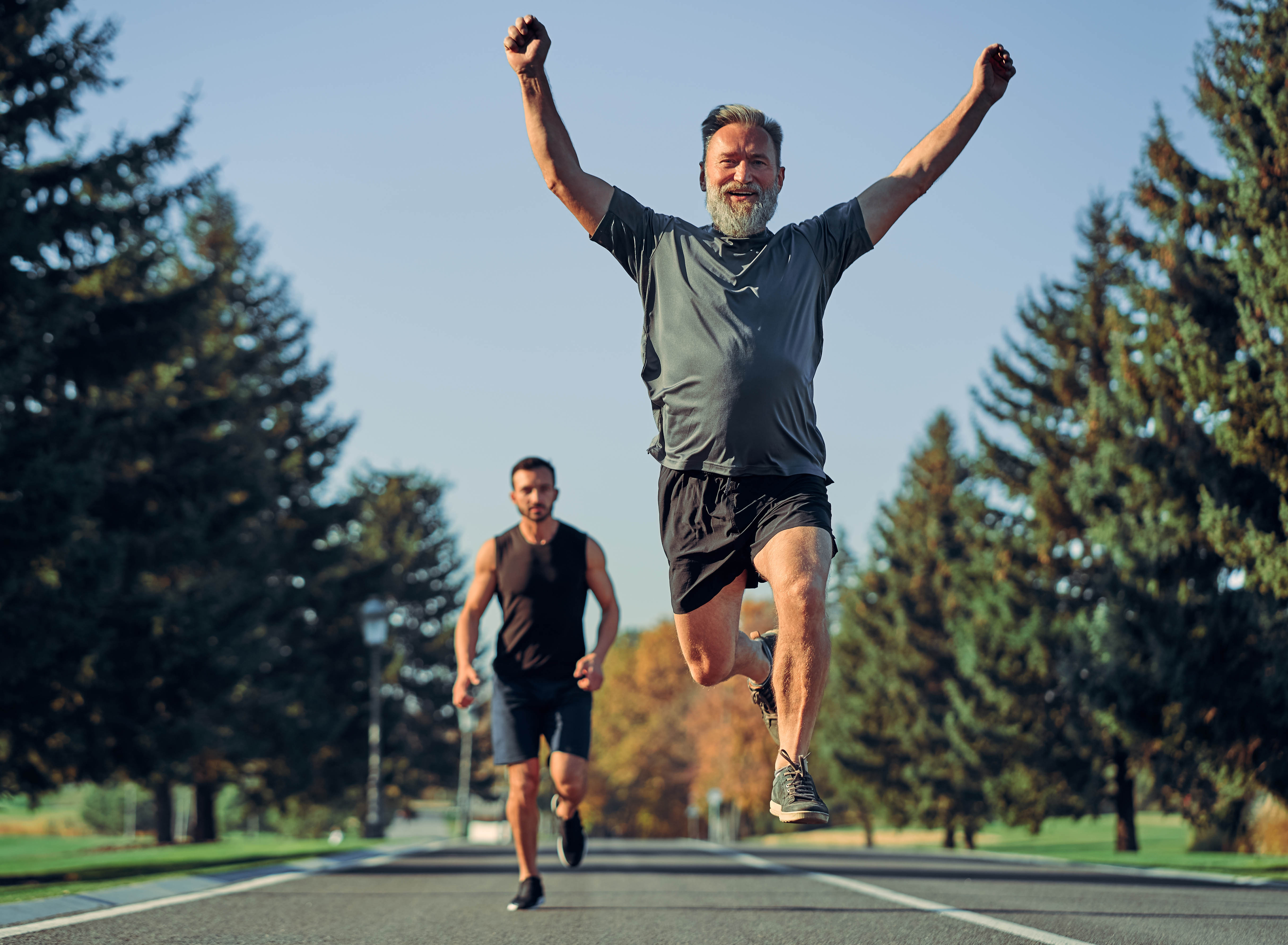 Older man crossing the finish line in a park with his arms raised and jumping into the air. Another, younger man is in the background running towards the older man.